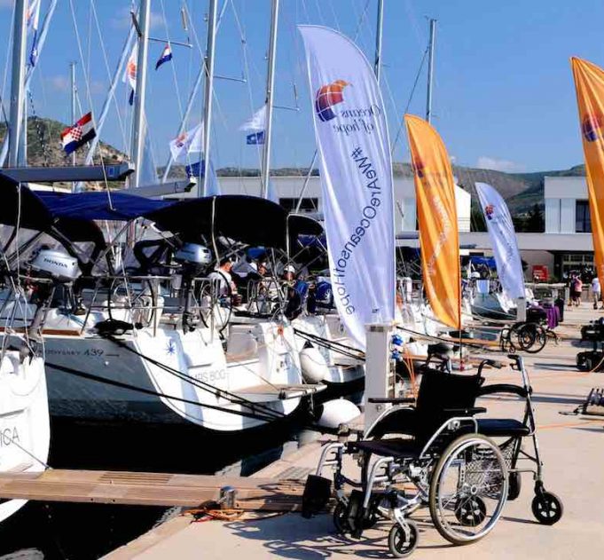 An empty wheelchair sits outside a yacht in the marina with blue skies and lots of other yachts in the background