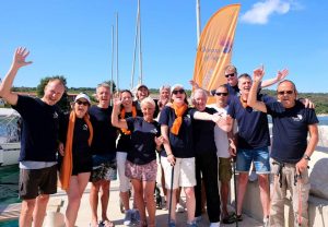 Crew of mixed ability sailors wearing Oceans of Hope blue tee shirts and waving and smiling in a marina with orange flag in the background