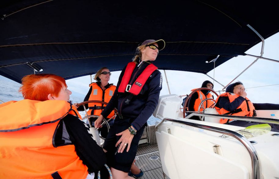 crew of five sailors in orange life jackets at the helm of a yacht under a black canopy