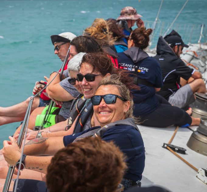 Sailors smiling and sitting with their legs over the side of a yacht with the ocean around them