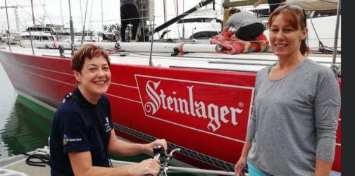 Ingrid and Gail pose in front of Steinlager 2, sitting on the pontoon in front of it. The red hull of the yacht and the name can be seen in the background.