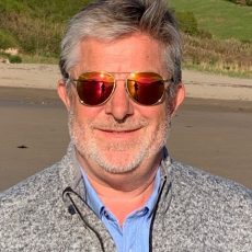 Headshot of David on a beach wearing sunglasses with a bronze reflection of the sea in his sunglasses
