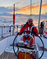 Heidi in a red sailing jacket at the helm of a yacht with a golden sunset over the waves in the background