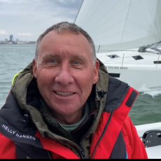 John wears a red sailing jacket and is smiling on the deck with a yacht sailing past in the background