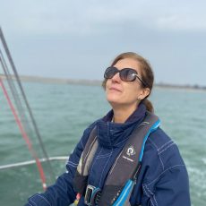 Kate is at the helm of a yacht wearing a blue sailing jacket with the sea in the background