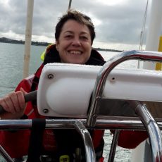 Ingrid is wearing a red sailing jacket and smiling at the helm of a yacht