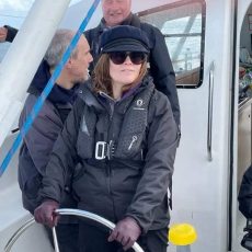 Lynwen is wearing a dark blue sailing jacket, sunglasses and hat at the helm of a yacht with two other sailors in the background