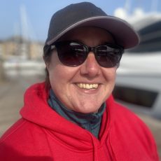 Headshot of Trish smiling wearing a red Oceans of Hope hoodie, sunglasses and a dark cap