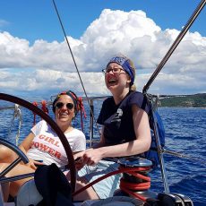 Image of Liz and Marina sitting at the helm of a yacht smiling and laughing with the ocean in the background